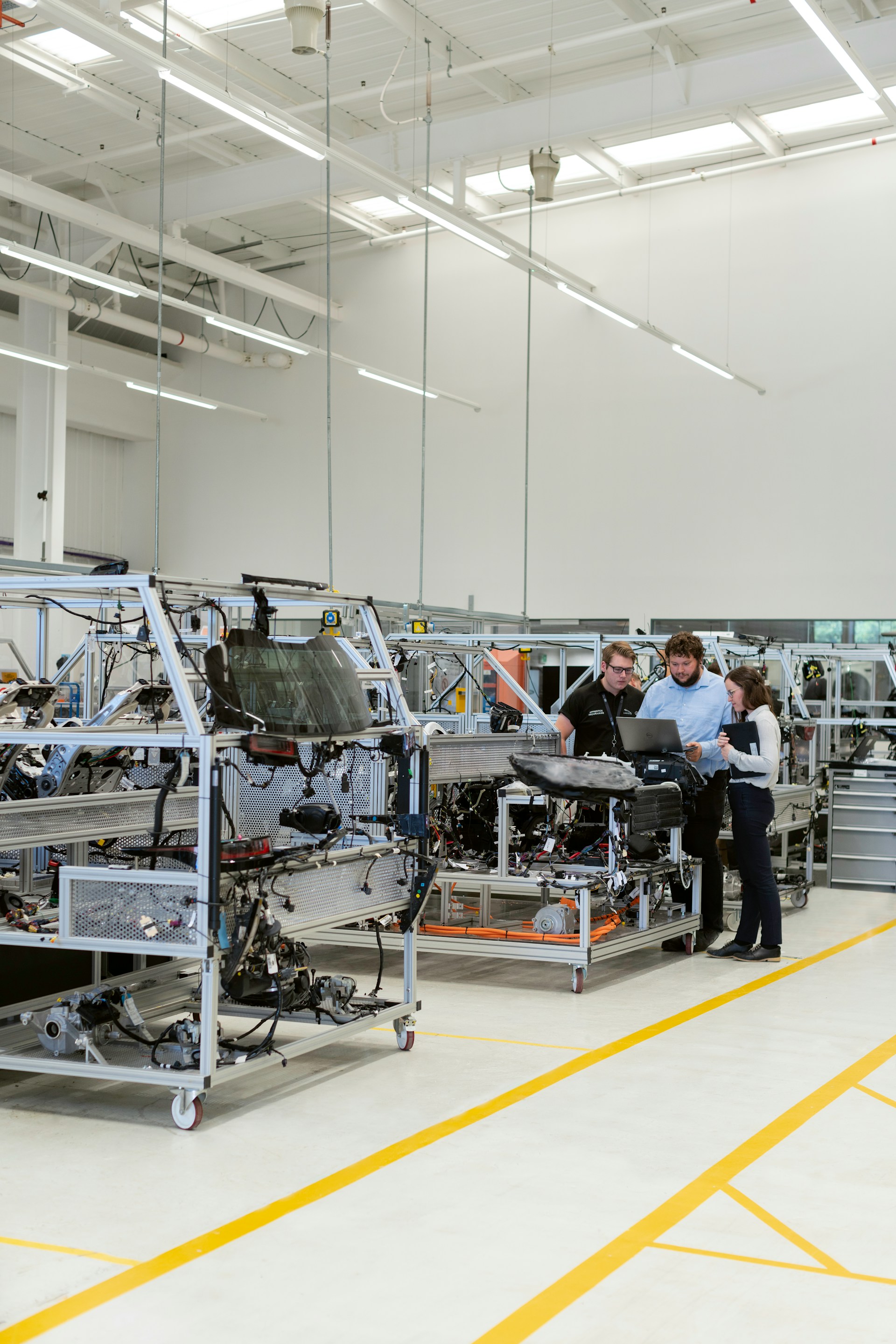 Three engineers review a laptop beside prototype vehicle assemblies on a plant floor