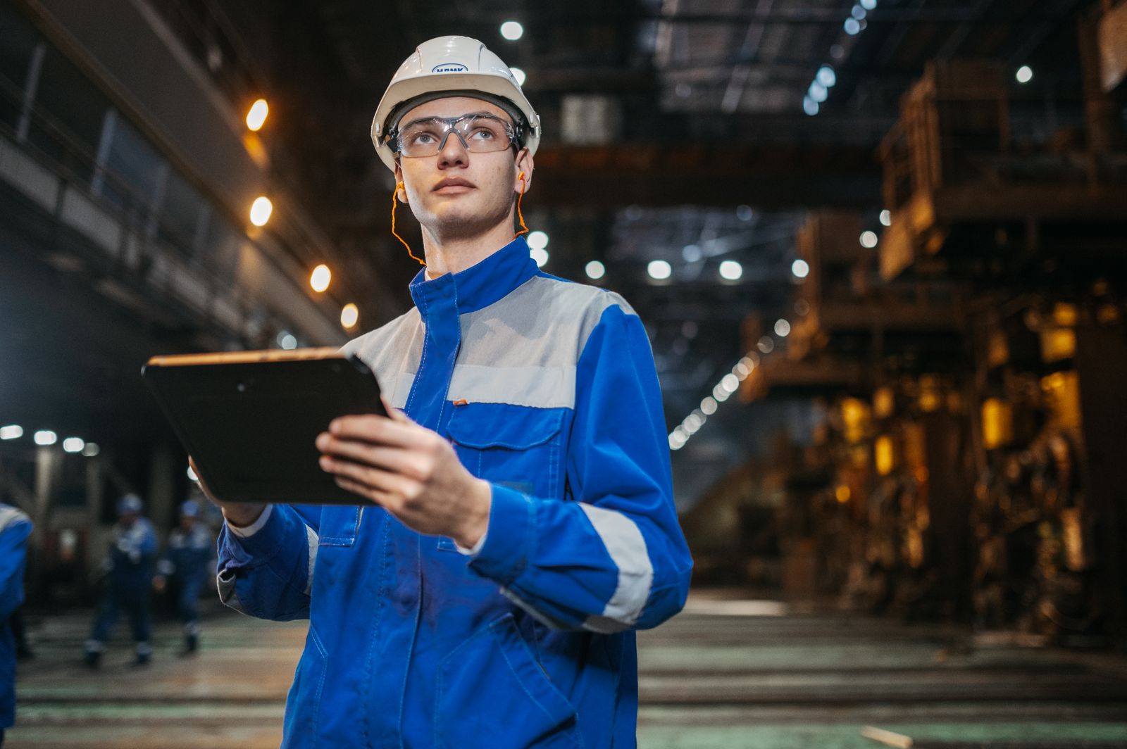 An engineer in safety gear uses a tablet in a large plant