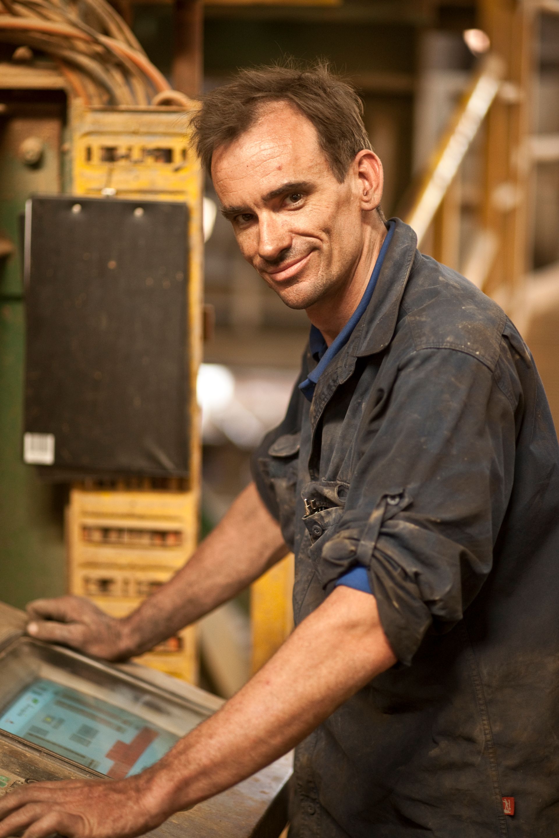 A smiling machine operator in safety glasses stands beside production equipment