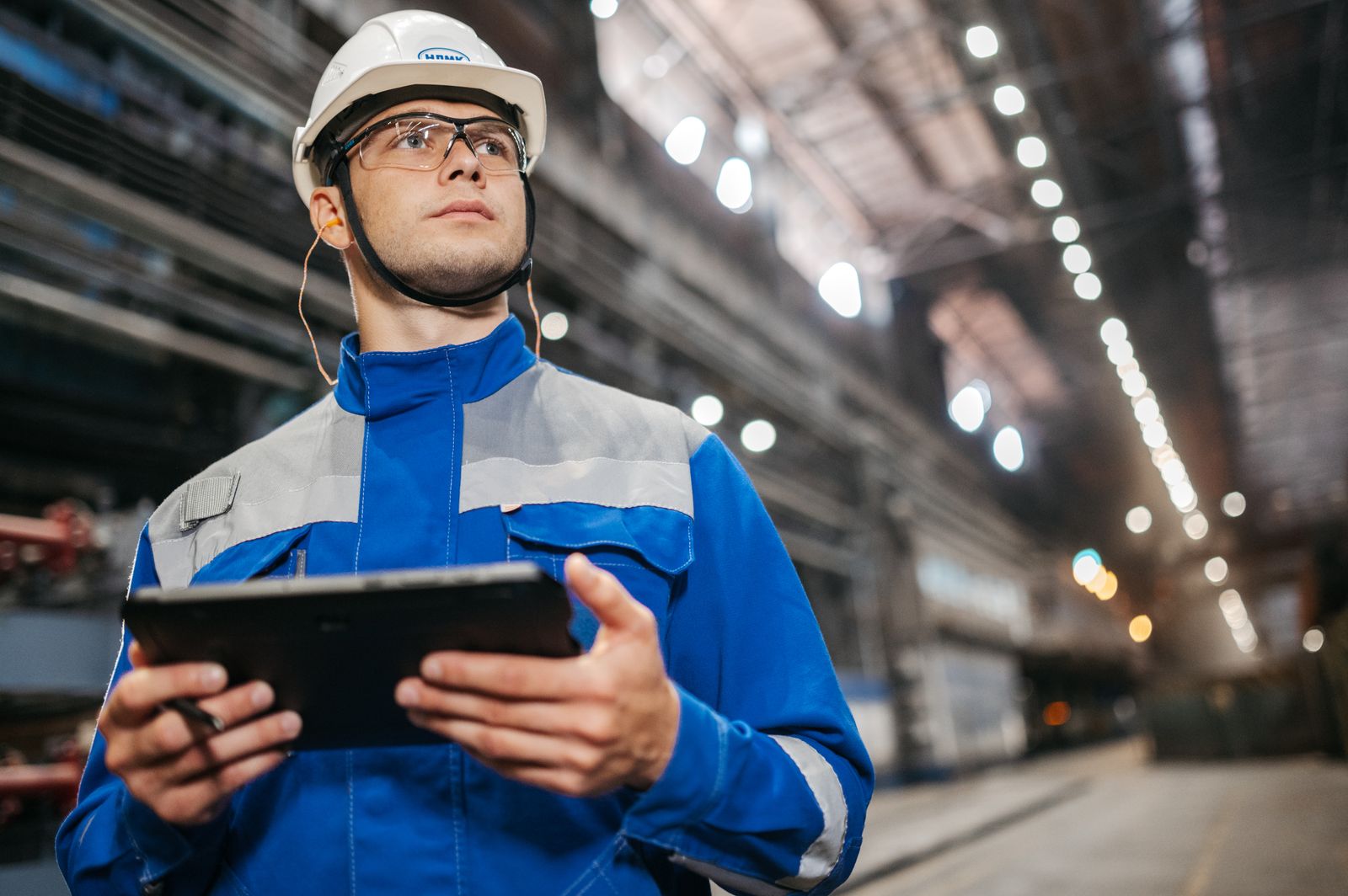 A factory worker in safety gear checks a tablet on the floor